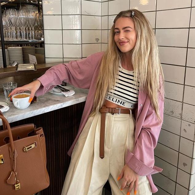 La influencer Emili Sindlev posa guiñando un ojo en una cafetería con el pelo liso, una camisa rosa y unos pantalones beige