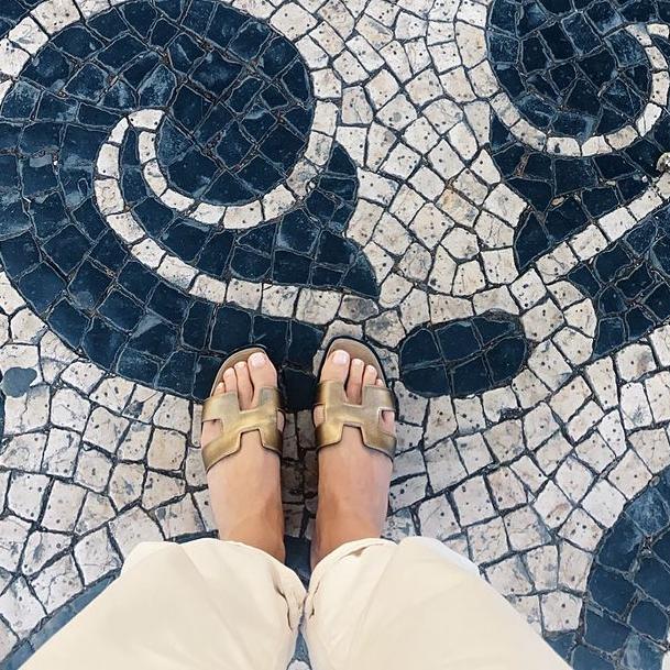 Los pies de una chica con sandalias doradas sobre un suelo de mosaico