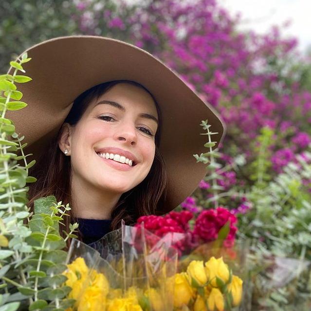 Anne Hathaway con un ramo de flores, sonriendo tras él con un sombrero y sin maquillaje