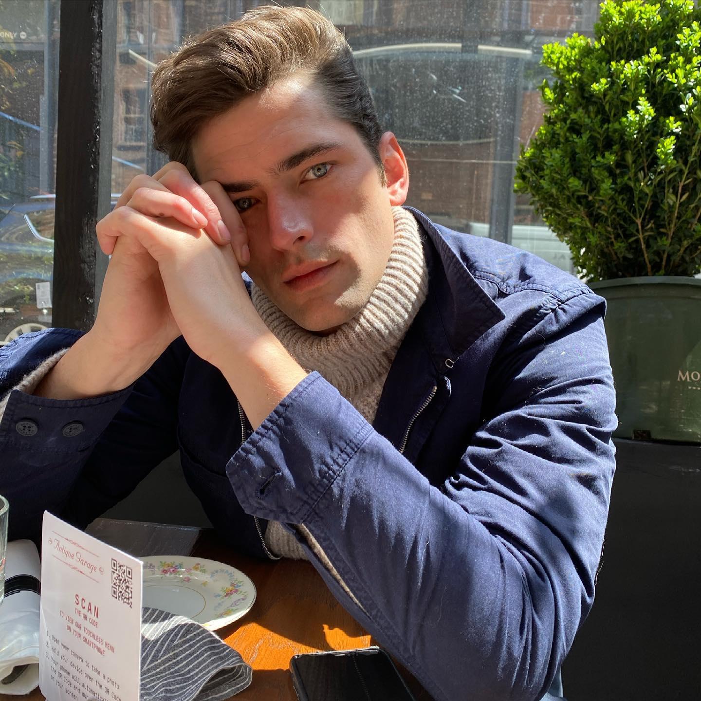 El modelo Sean O'Pry posa en una terraza co una chaqueta azul y un jersey beige apoyando un lado de la cara sobre las manos
