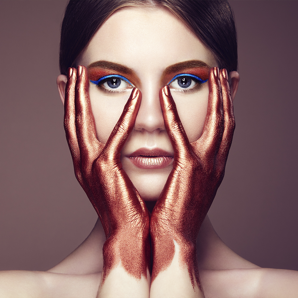 Imagen de una mujer con las manos maquilladas en color bronce sobre la cara con los ojos maquillados en color bronce y un eyeliner azul