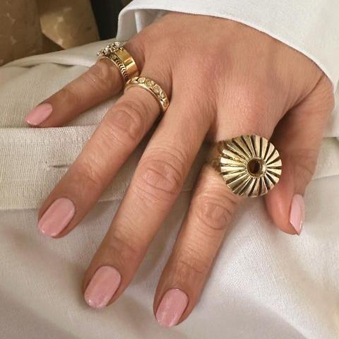 Imagen de la mano de Jennifer Lopez con una manicura en tonos rosa claro y tres maxi anillos dorados